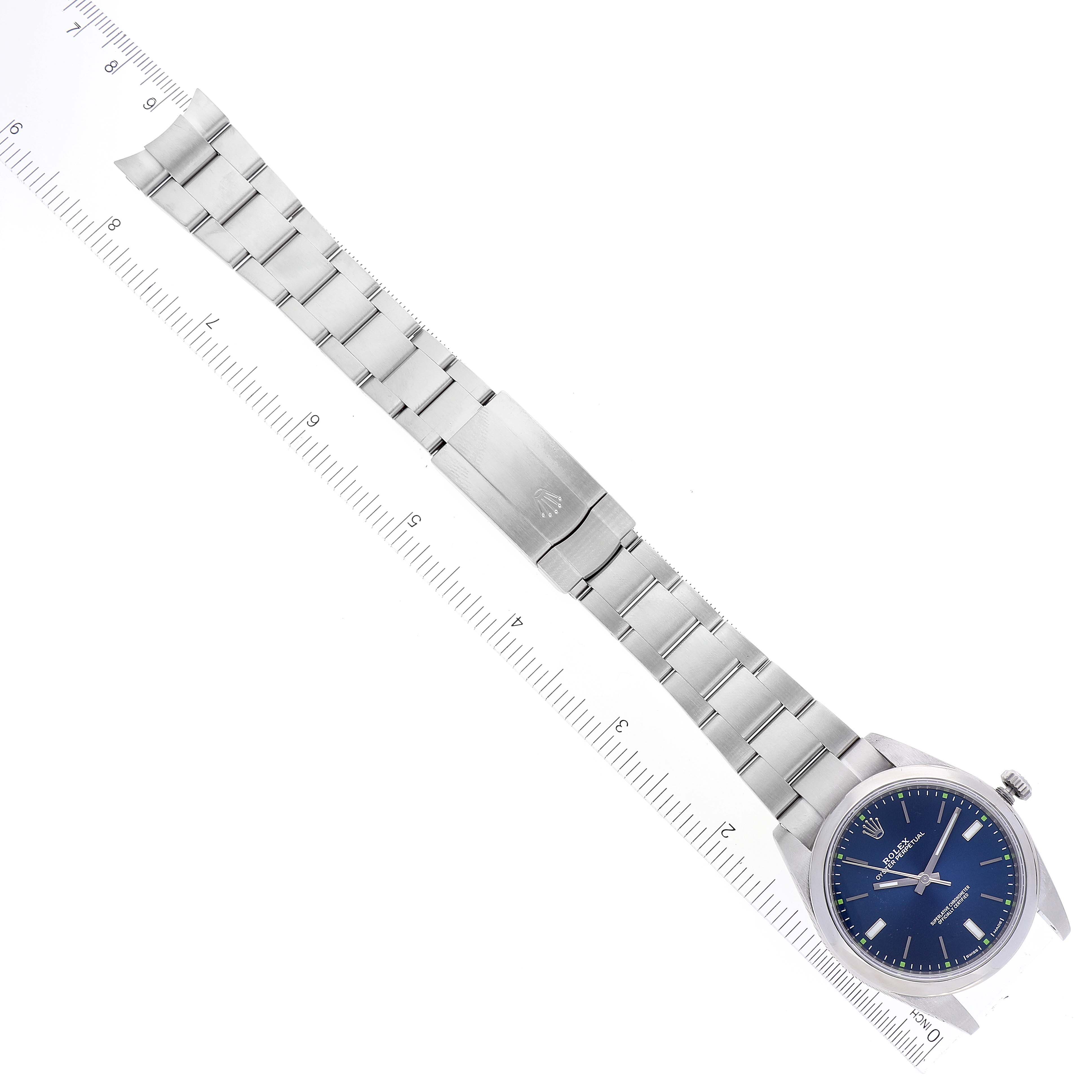 This image displays the Rolex Oyster Perpetual 114300 Men's Stainless Steel Blue Dial 114300 Men's Stainless Steel Blue Dial watch lying flat, showing the entire watch face, bracelet, and clasp against a ruler for scale.