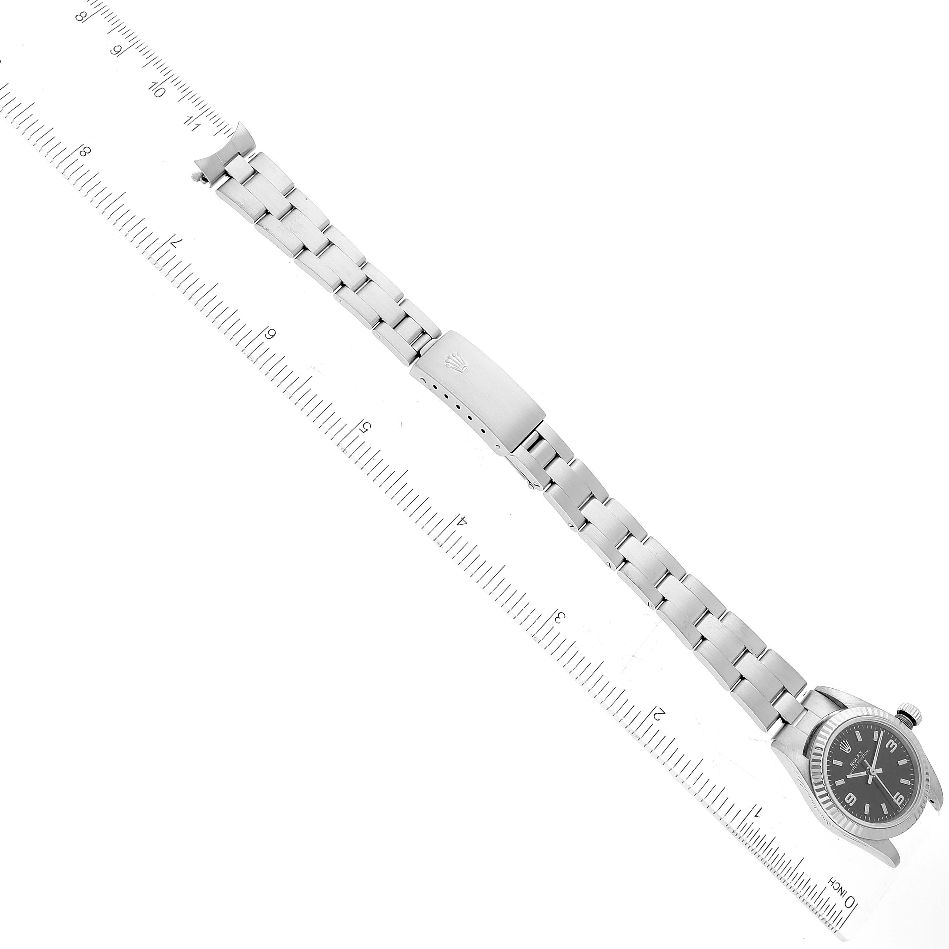 The image shows the top view of a Rolex Oyster Perpetual 76094 Women's Stainless Steel Black Dial 76094 Women's Stainless Steel Black Dial watch, highlighting its face, bracelet, and clasp next to a ruler for scale.