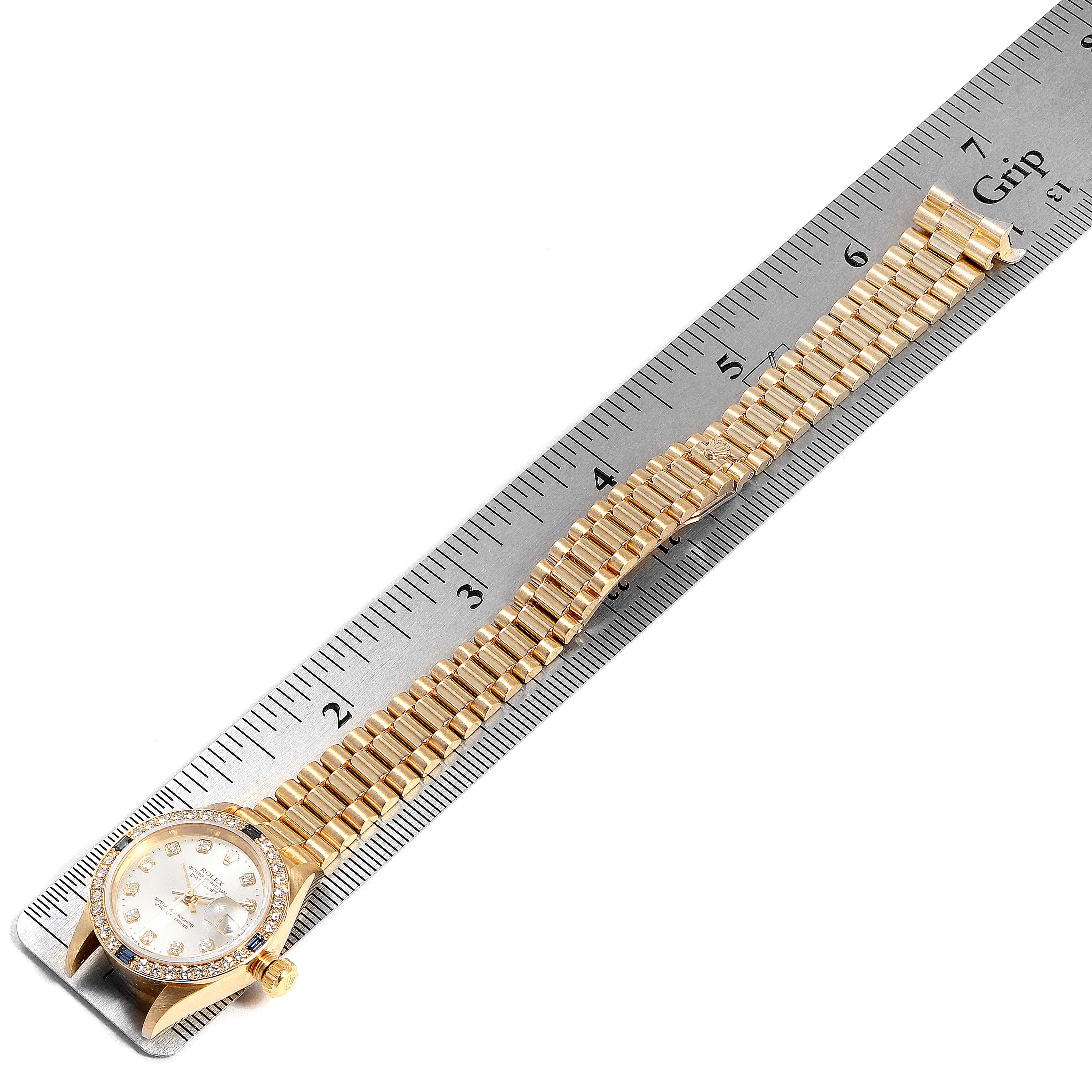 The image shows a gold Rolex President 79088 Women
s Yellow Gold Silver Dial 79088 Women
s Yellow Gold Silver Dial model watch placed on a ruler, highlighting its bracelet and face.