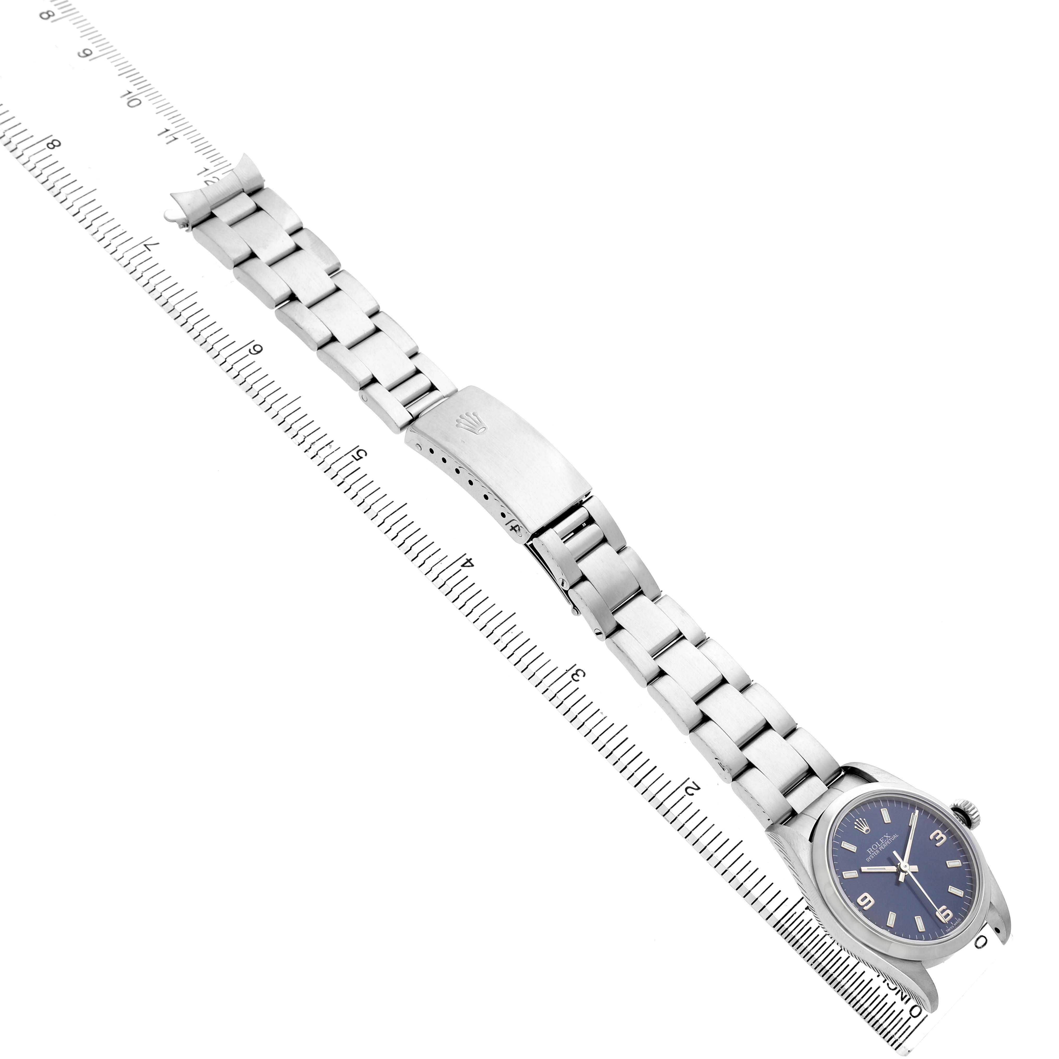 The Rolex Oyster Perpetual 77080 Women's Stainless Steel Blue Dial watch is shown at an angle above with the full bracelet and clasp visible alongside a ruler.