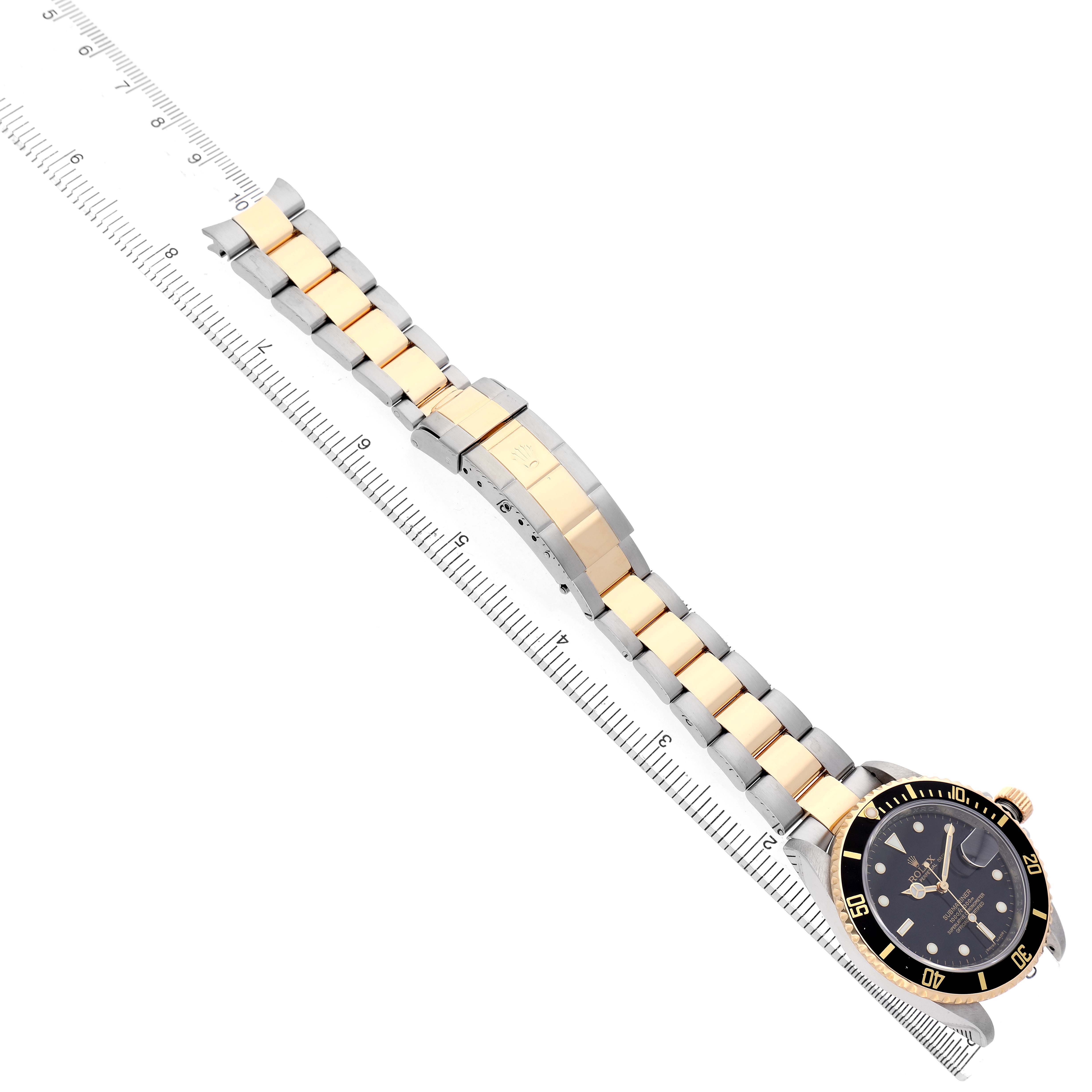 The image shows a Rolex Submariner 16613 Men's Steel and Gold (two tone) Black Dial watch with a two-tone gold and silver bracelet, placed diagonally next to a ruler.