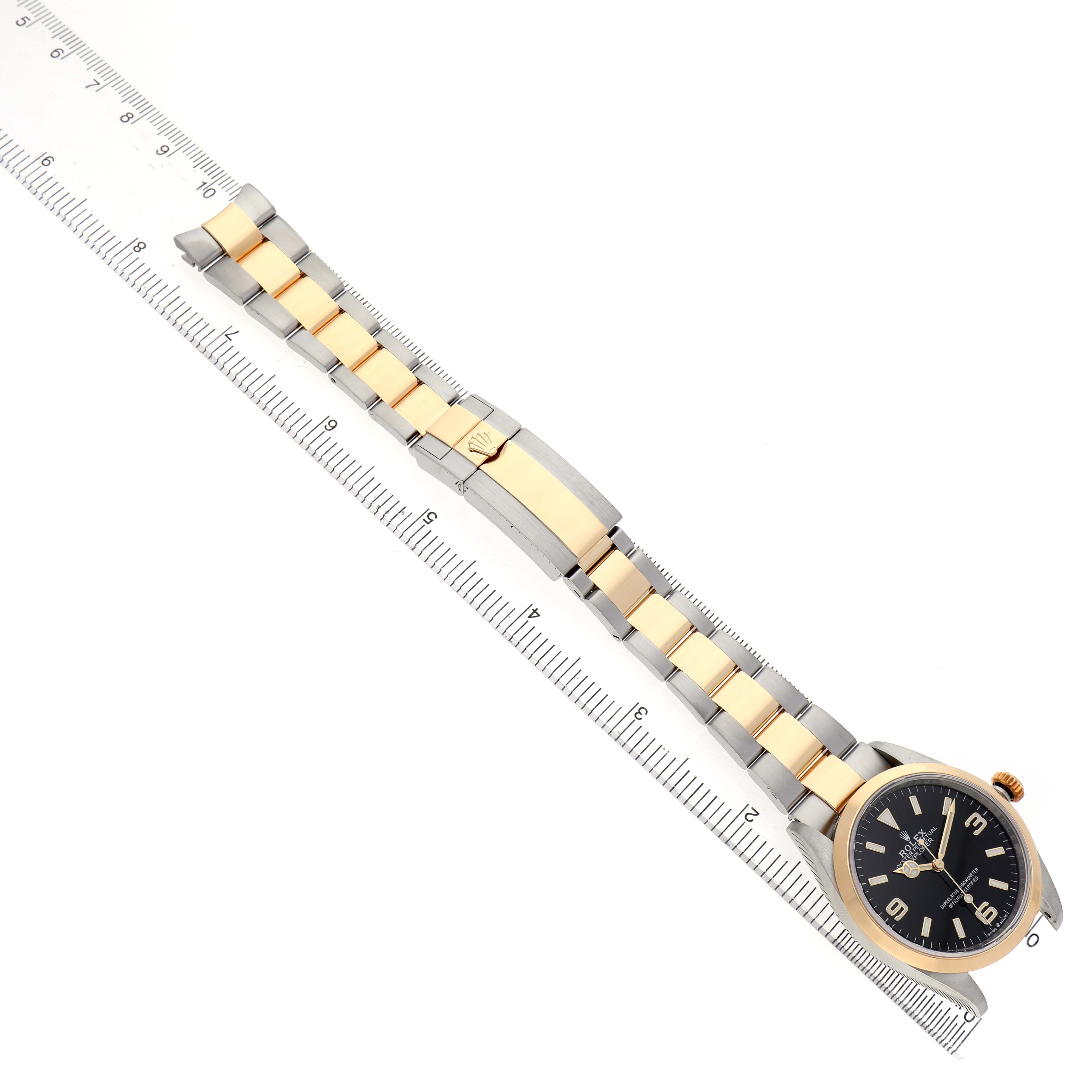 The Rolex Explorer 124273 Men's Steel and Gold (two tone) Black Dial watch is shown from a top-down angle, highlighting its two-tone bracelet and clasp alongside a ruler.