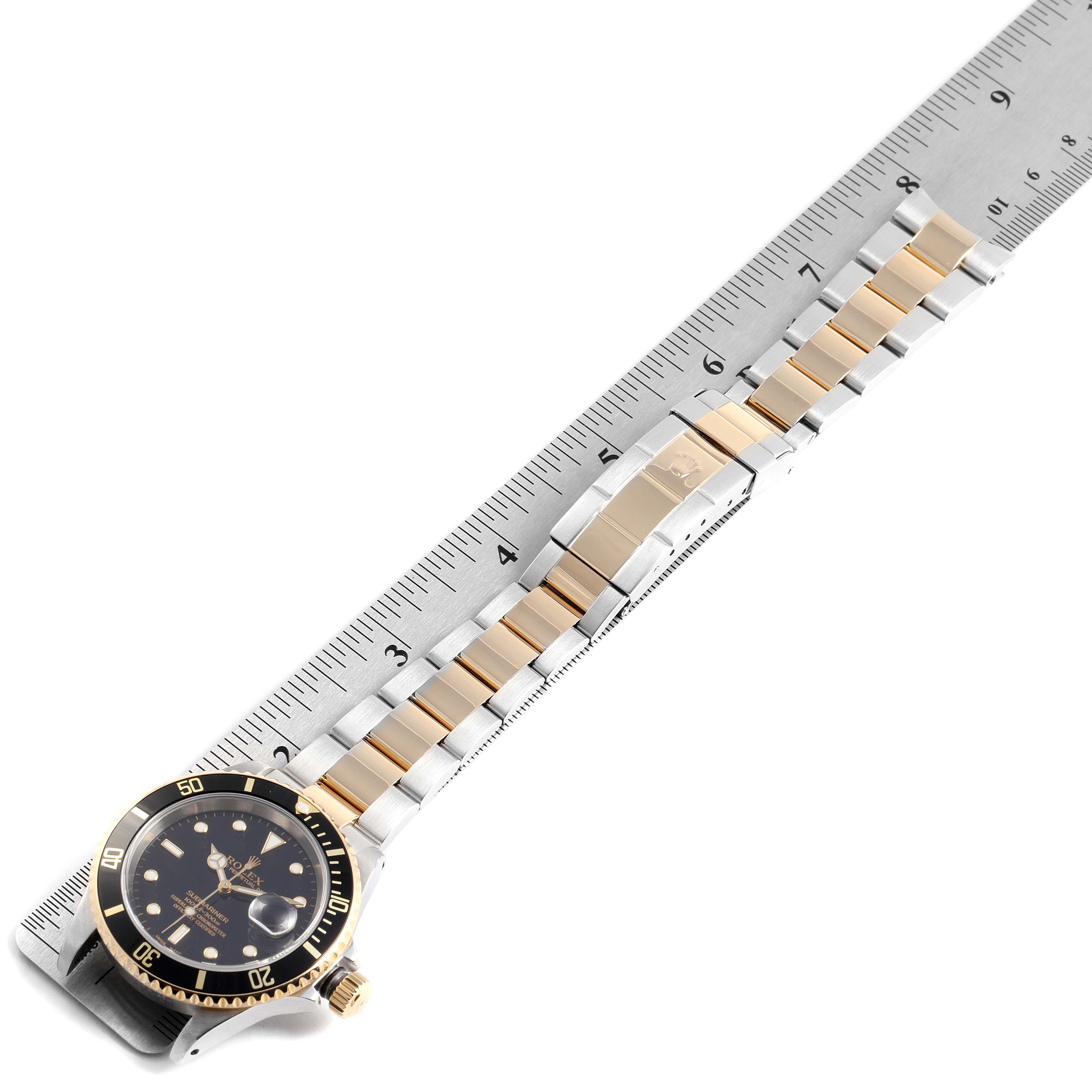 The Rolex Submariner 16613 Men
s Steel and Gold (two tone) Black Dial 16613 Men
s Steel and Gold (two tone) Black Dial watch is shown from an overhead angle, displaying the face, bezel, and full bracelet alongside a ruler.