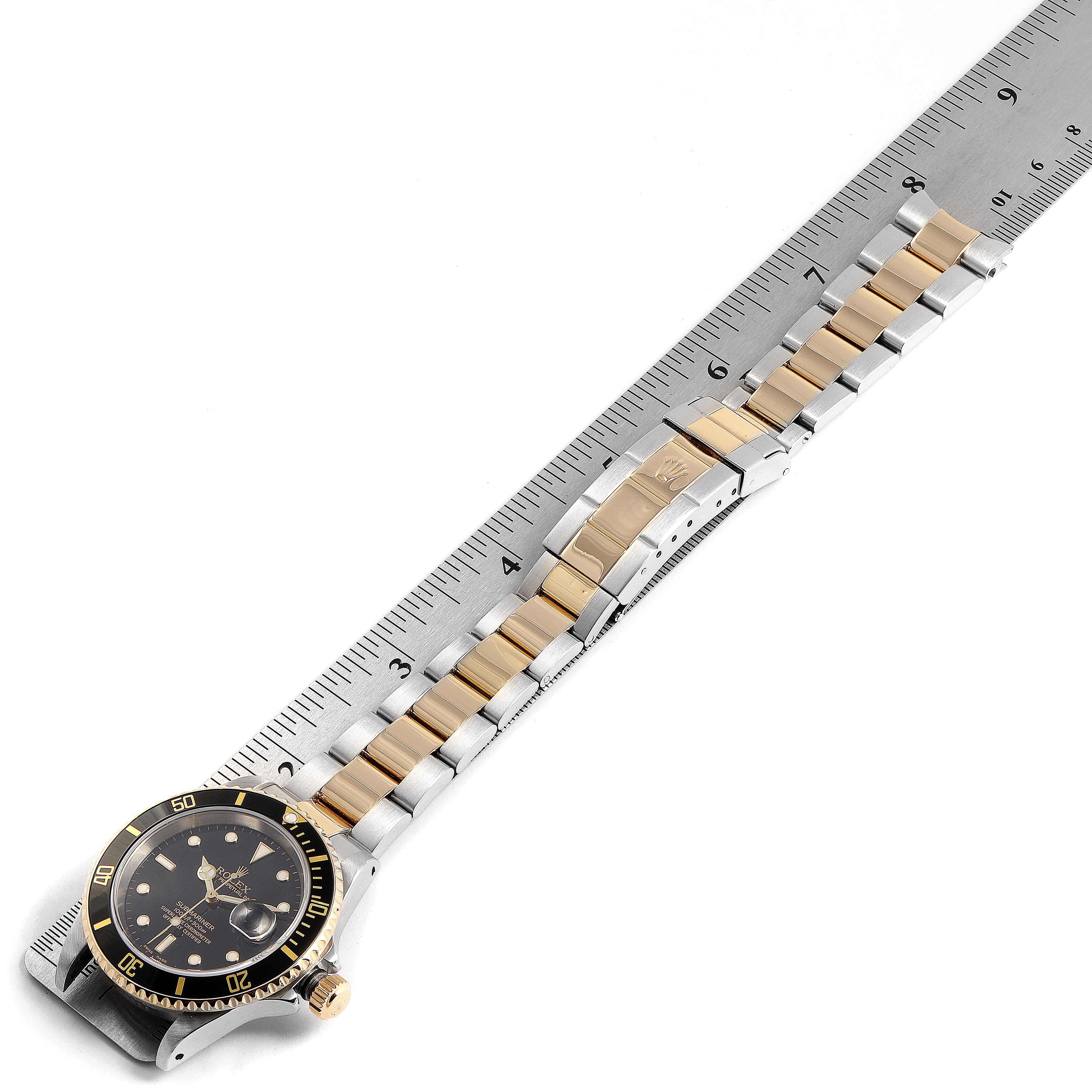 The image shows a Rolex Submariner 16613 Men
s Steel and Gold (two tone) Black Dial 16613 Men
s Steel and Gold (two tone) Black Dial watch laid flat on a ruler, displaying its face, bezel, crown, and the full bracelet.