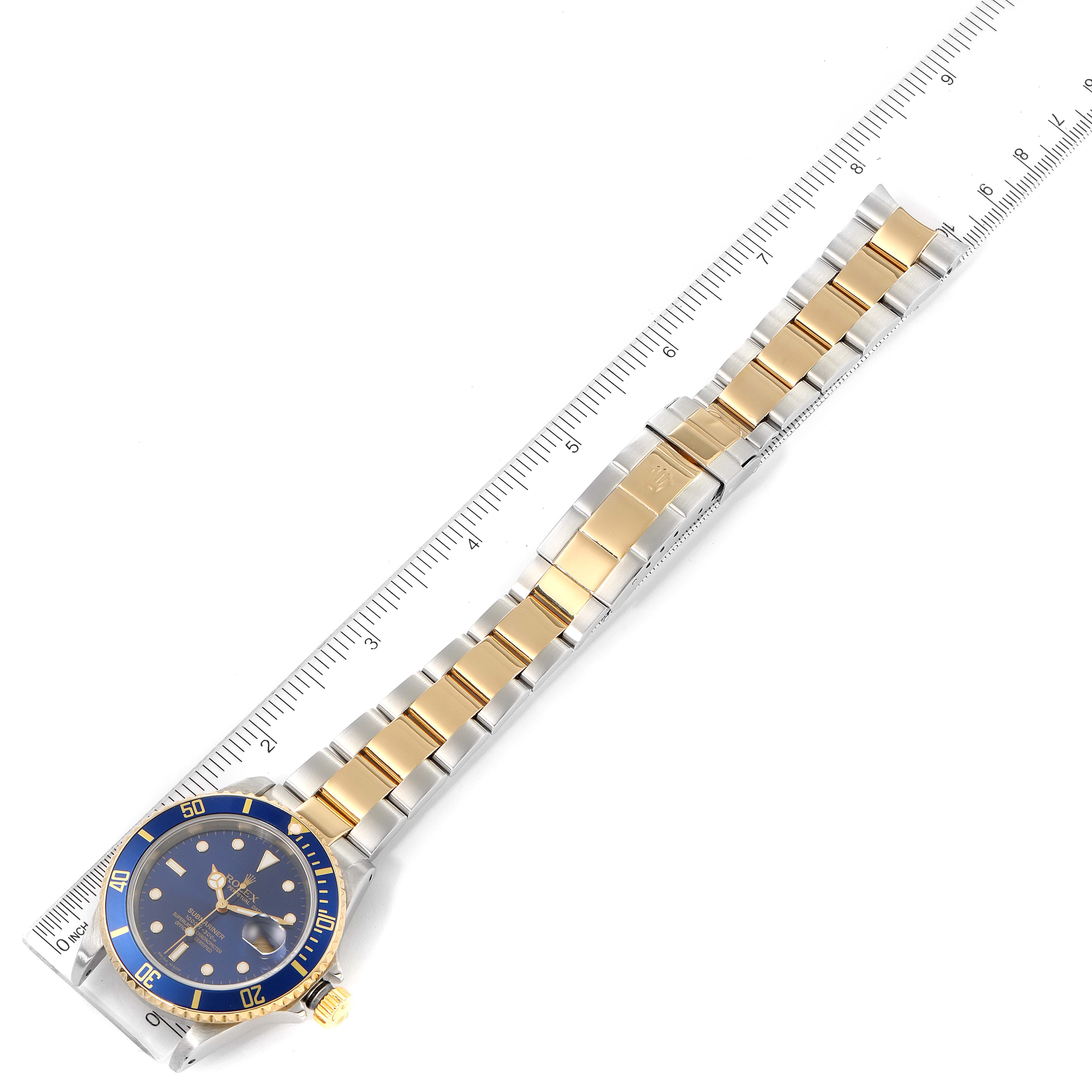 The Rolex Submariner 16613 Men's Steel and Gold (two tone) Blue Dial 16613 Men's Steel and Gold (two tone) Blue Dial watch is shown laid flat with both the face and bracelet visible, next to a ruler for scale.