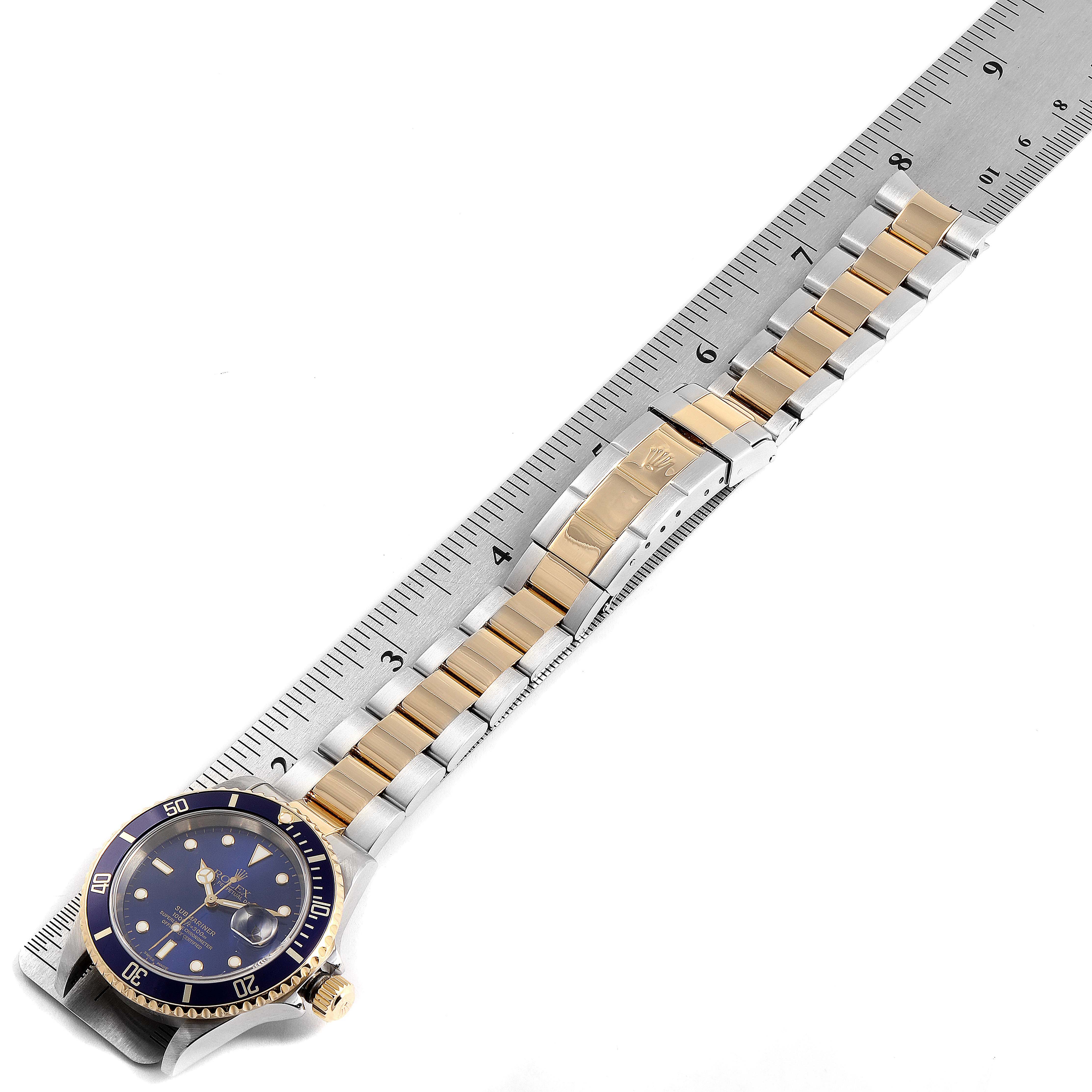 The image shows a Rolex Submariner 16613 Men
s Steel and Gold (two tone) Blue Dial 16613 Men
s Steel and Gold (two tone) Blue Dial watch placed next to a ruler, highlighting its face and two-tone metal bracelet.