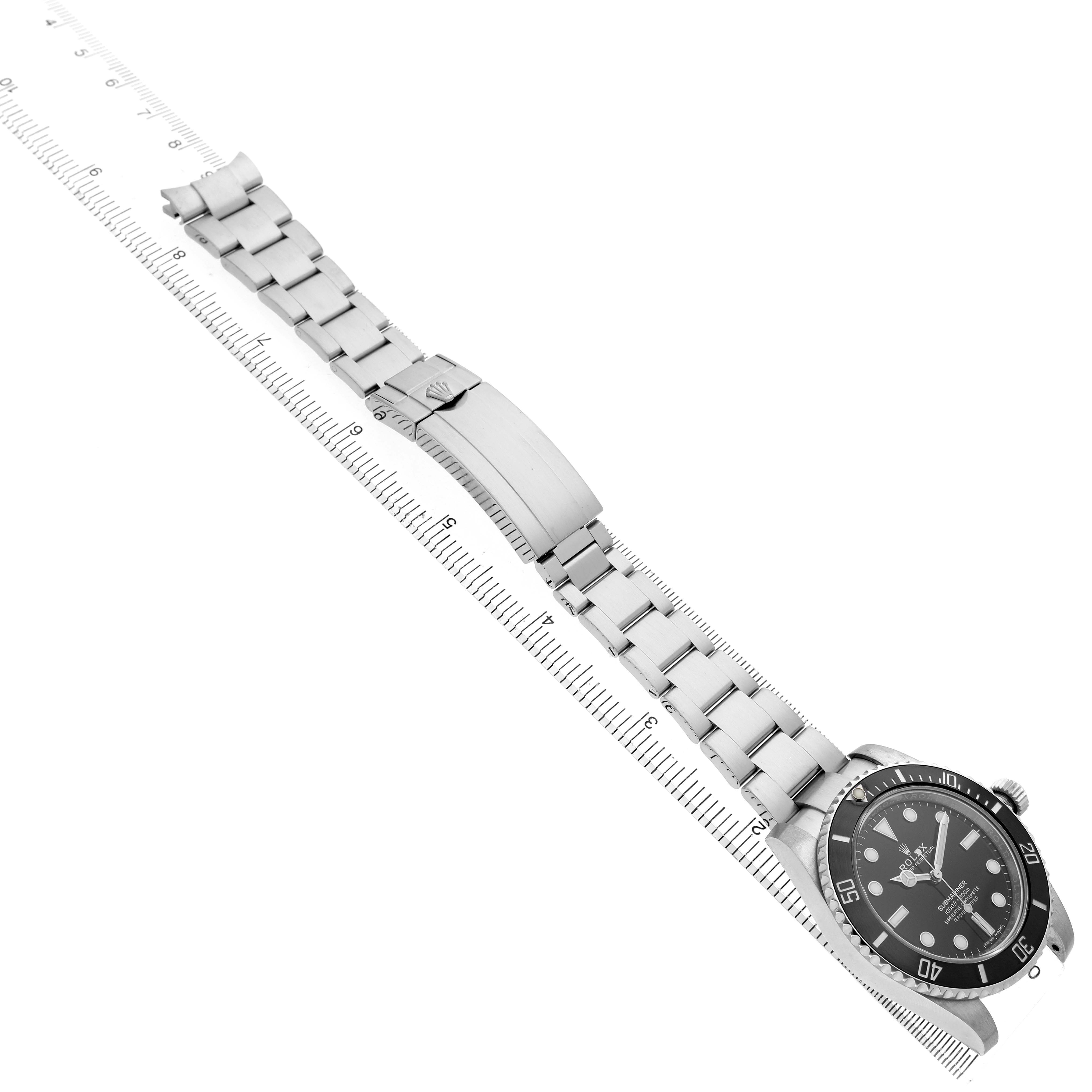 The Rolex Submariner 114060 Men's Stainless Steel Black Dial watch is shown diagonally, displaying the face, bezel, and full metal bracelet with a closed clasp next to a ruler.