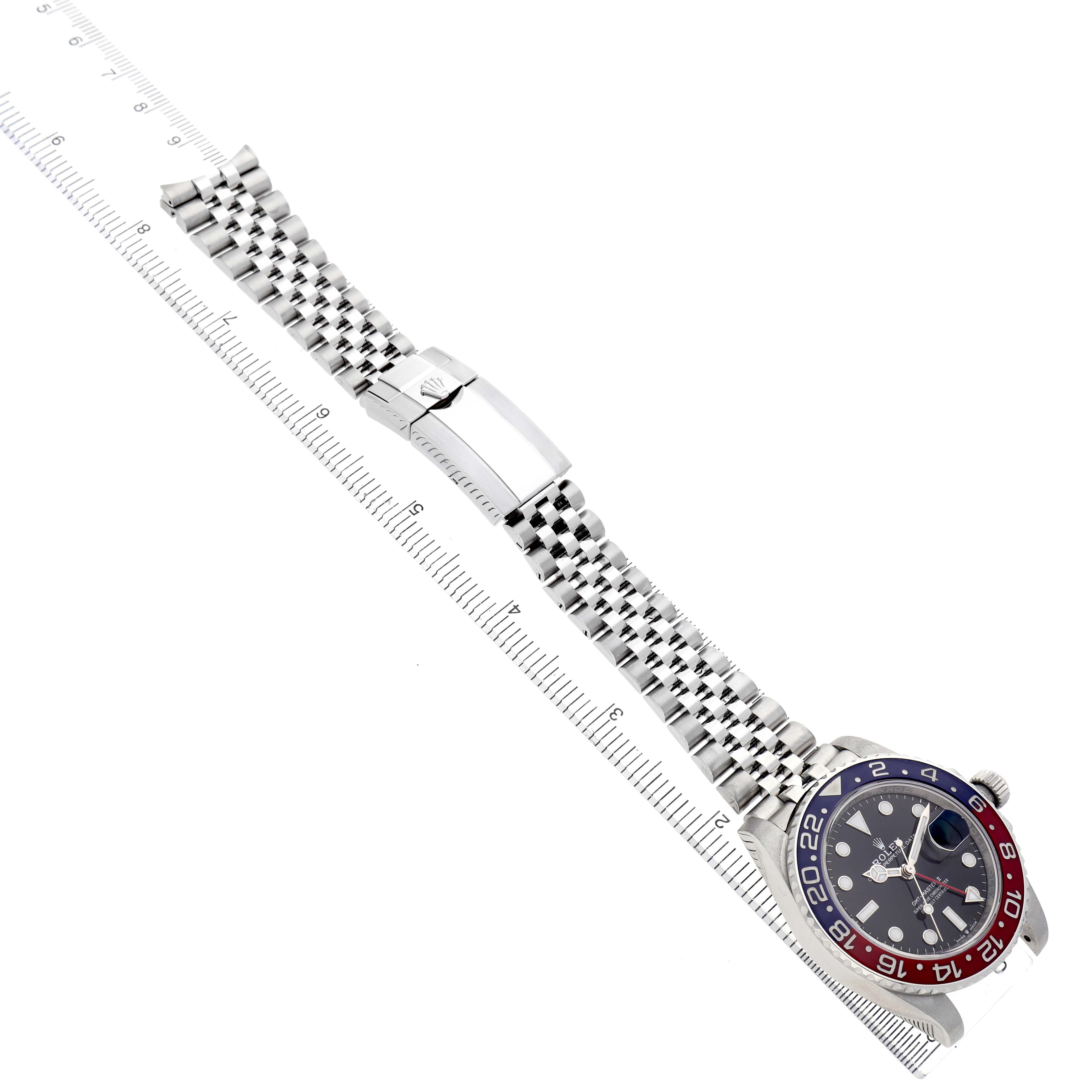 The Rolex GMT-Master 126710BLRO Men's Stainless Steel Black Dial watch is shown from an overhead angle, featuring the bezel, bracelet, and clasp alongside a ruler.