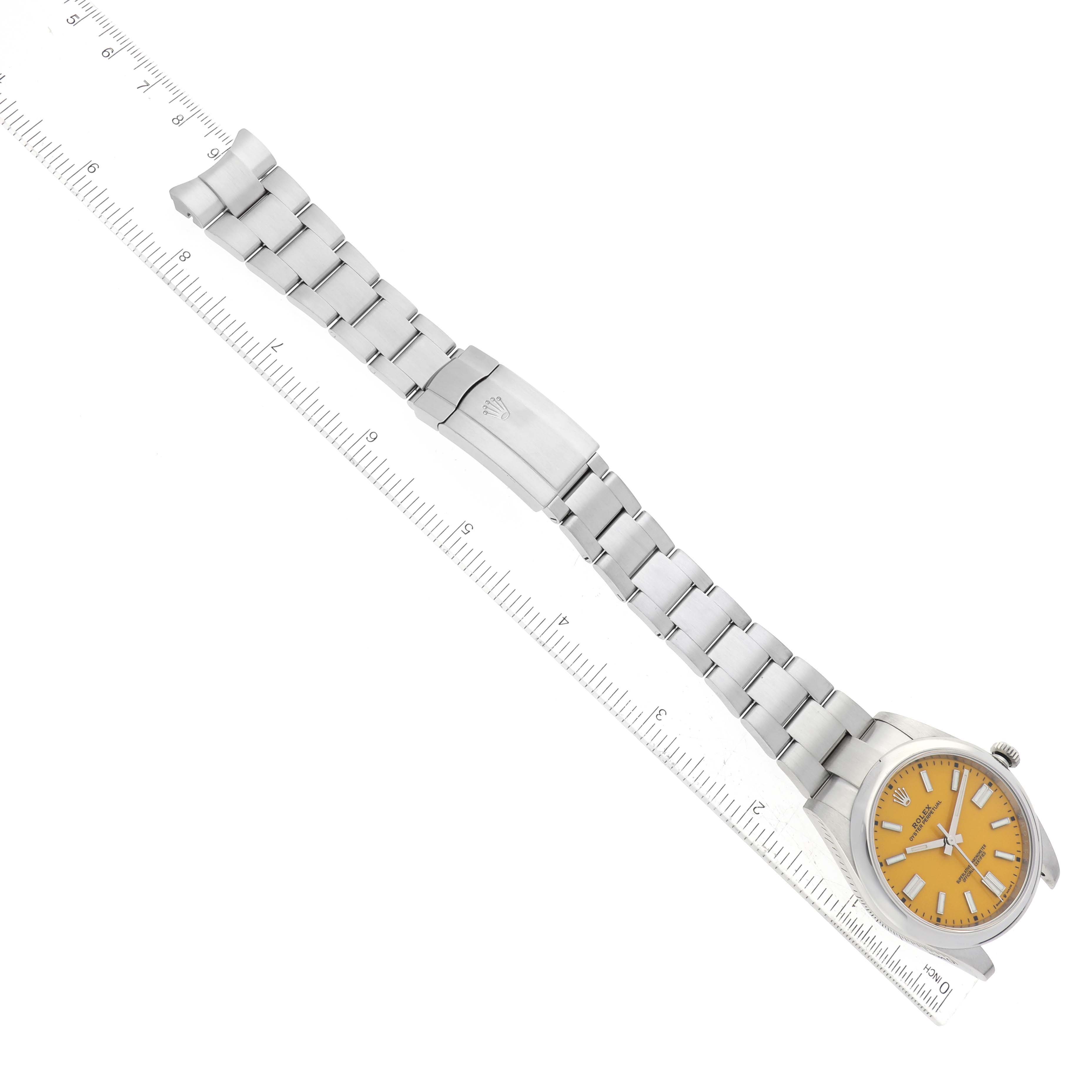 The image shows a Rolex Oyster Perpetual 124300 Men
s Stainless Steel Yellow Dial 124300 Men
s Stainless Steel Yellow Dial watch with an orange dial, laid flat with a full view of the metal bracelet and clasp along a ruler.
