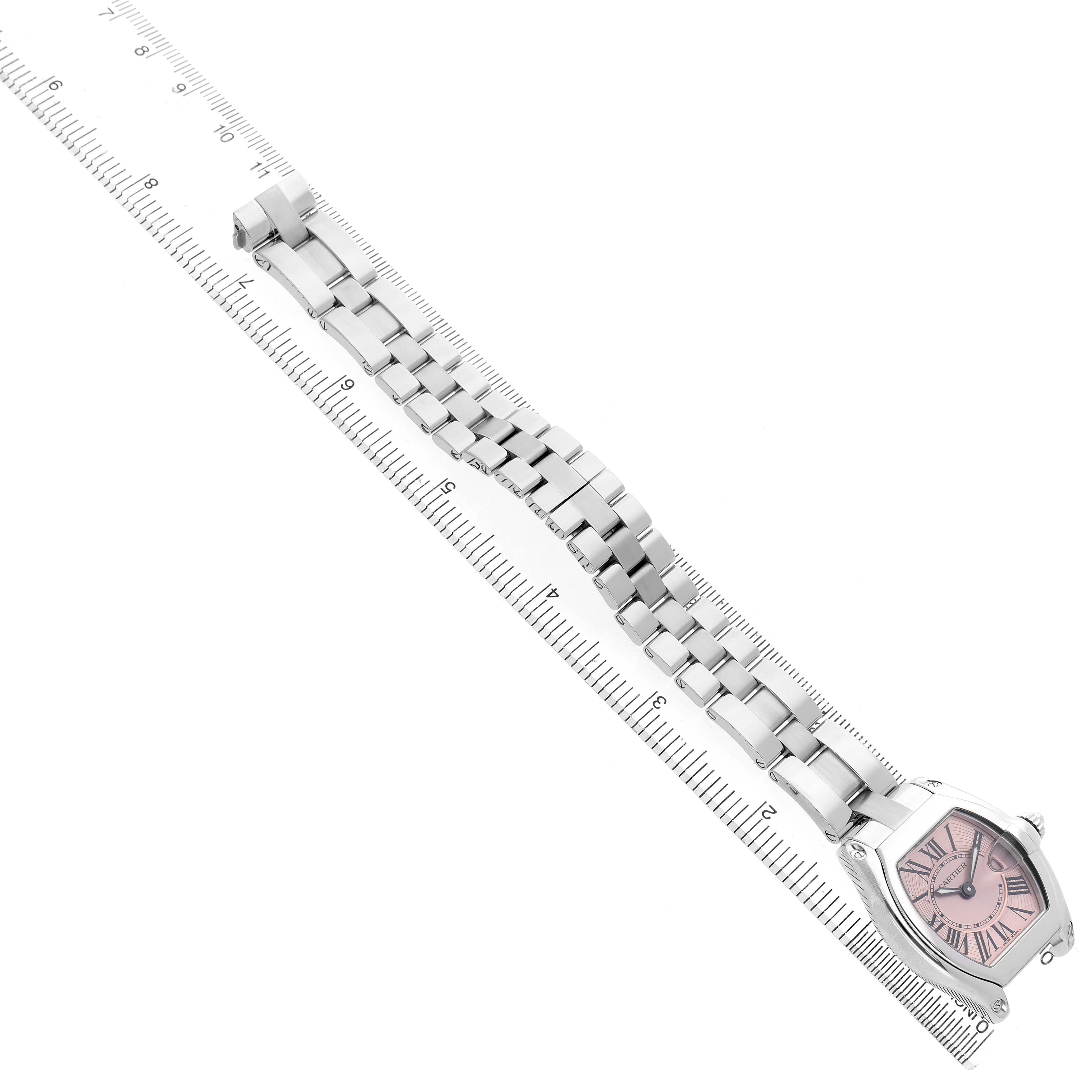The Cartier Roadster W62017V3 Women's Stainless Steel Pink Dial watch is shown at an angled top view, featuring a pink dial and metal bracelet laid over a ruler.