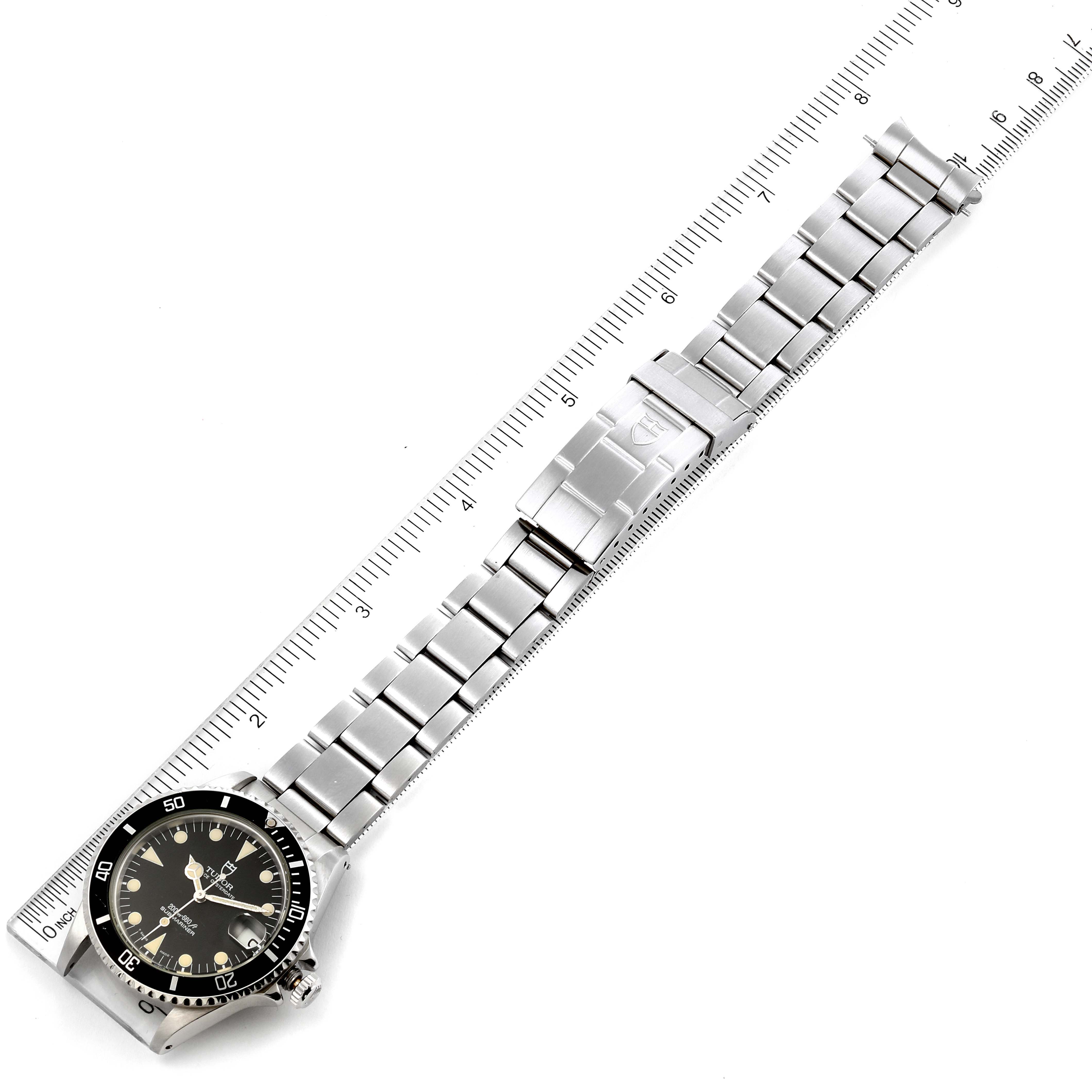 The image shows a Tudor Submariner 75090 Men
s Stainless Steel Black Dial 75090 Men
s Stainless Steel Black Dial watch laid flat with an angled view of its bracelet and clasp, alongside a ruler for size reference.