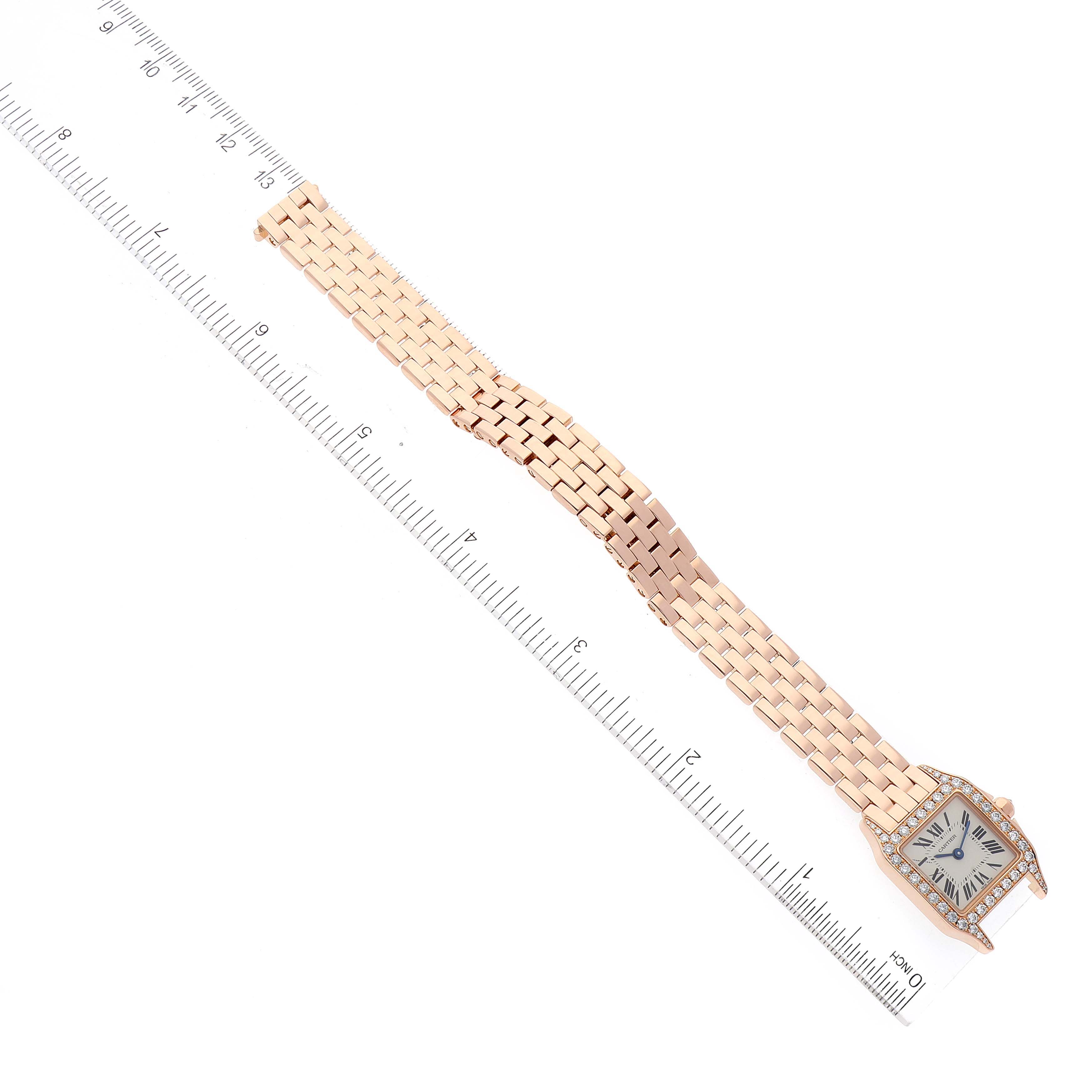 The Cartier Santos Demoiselle WF9011Z8 Women's Rose Gold Silver Dial WF9011Z8 Women's Rose Gold Silver Dial watch is shown in a flat lay angle, displaying the full watch length with its face and bracelet alongside a ruler.