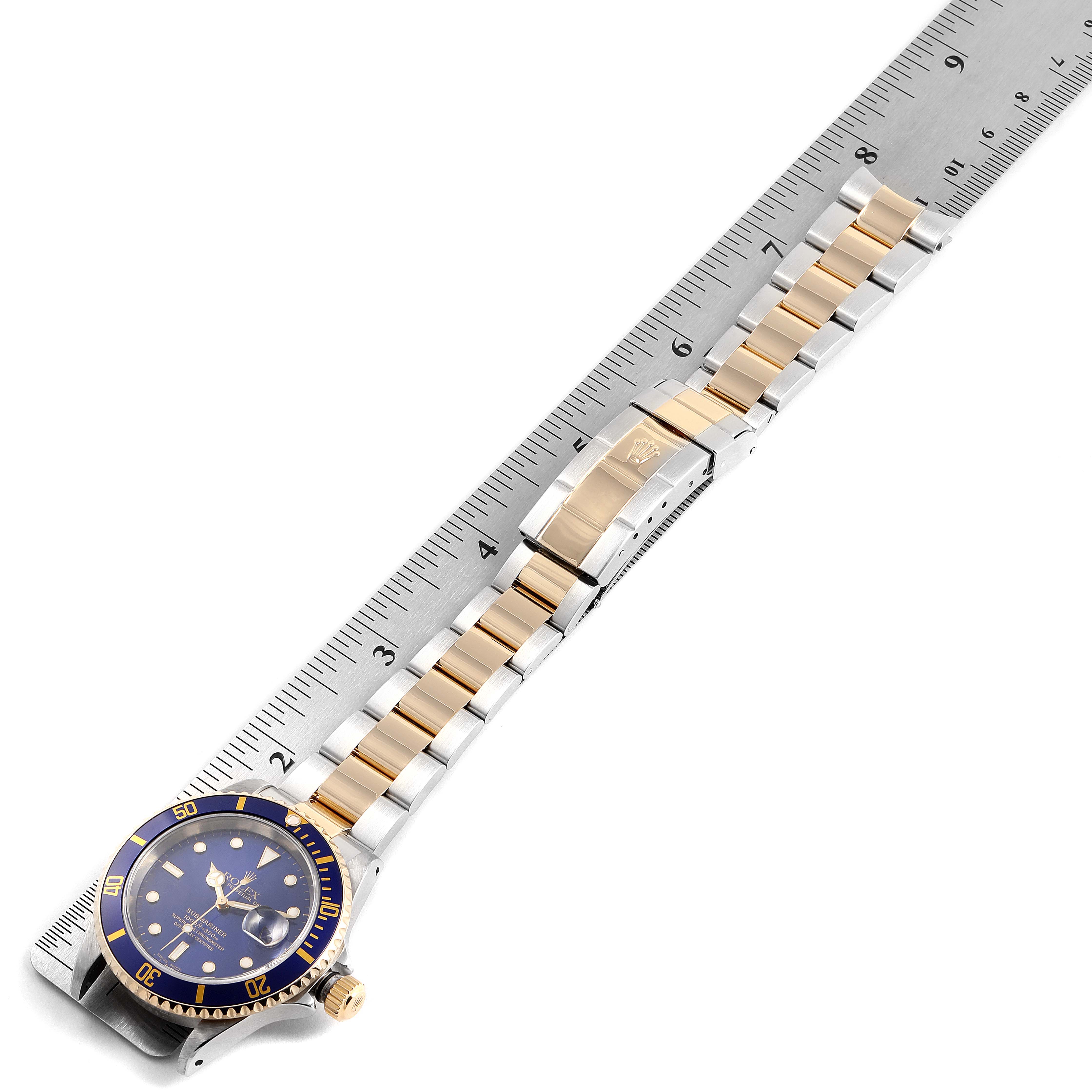 The image shows a Rolex Submariner 16613 Men's Steel and Gold (two tone) Blue Dial 16613 Men's Steel and Gold (two tone) Blue Dial watch laid flat next to a ruler, highlighting the strap and face.