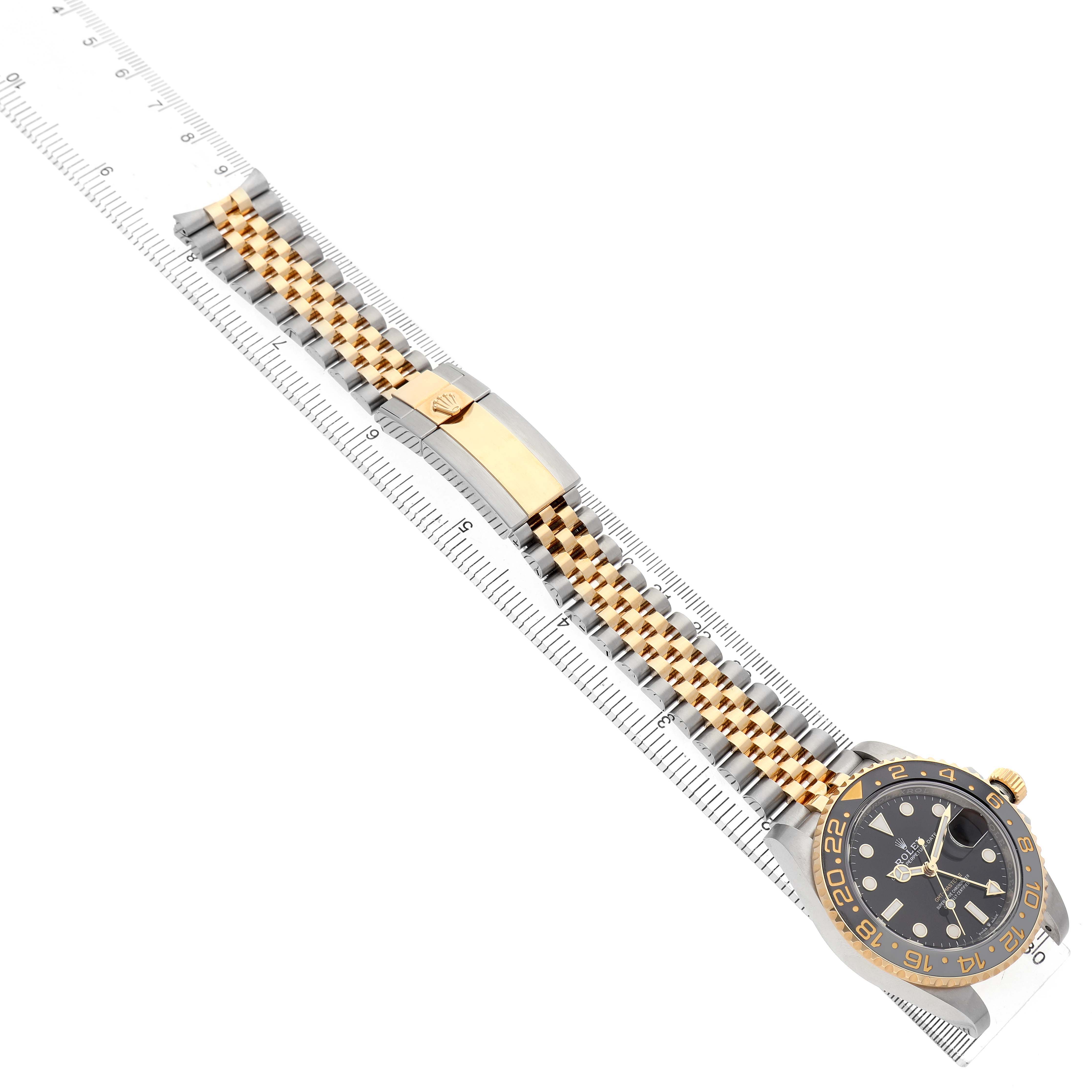 The Rolex GMT-Master 126713GRNR Men's Steel and Gold (two tone) Black Dial watch is shown at an angle revealing the bezel, face, clasp, and two-tone bracelet alongside a ruler.