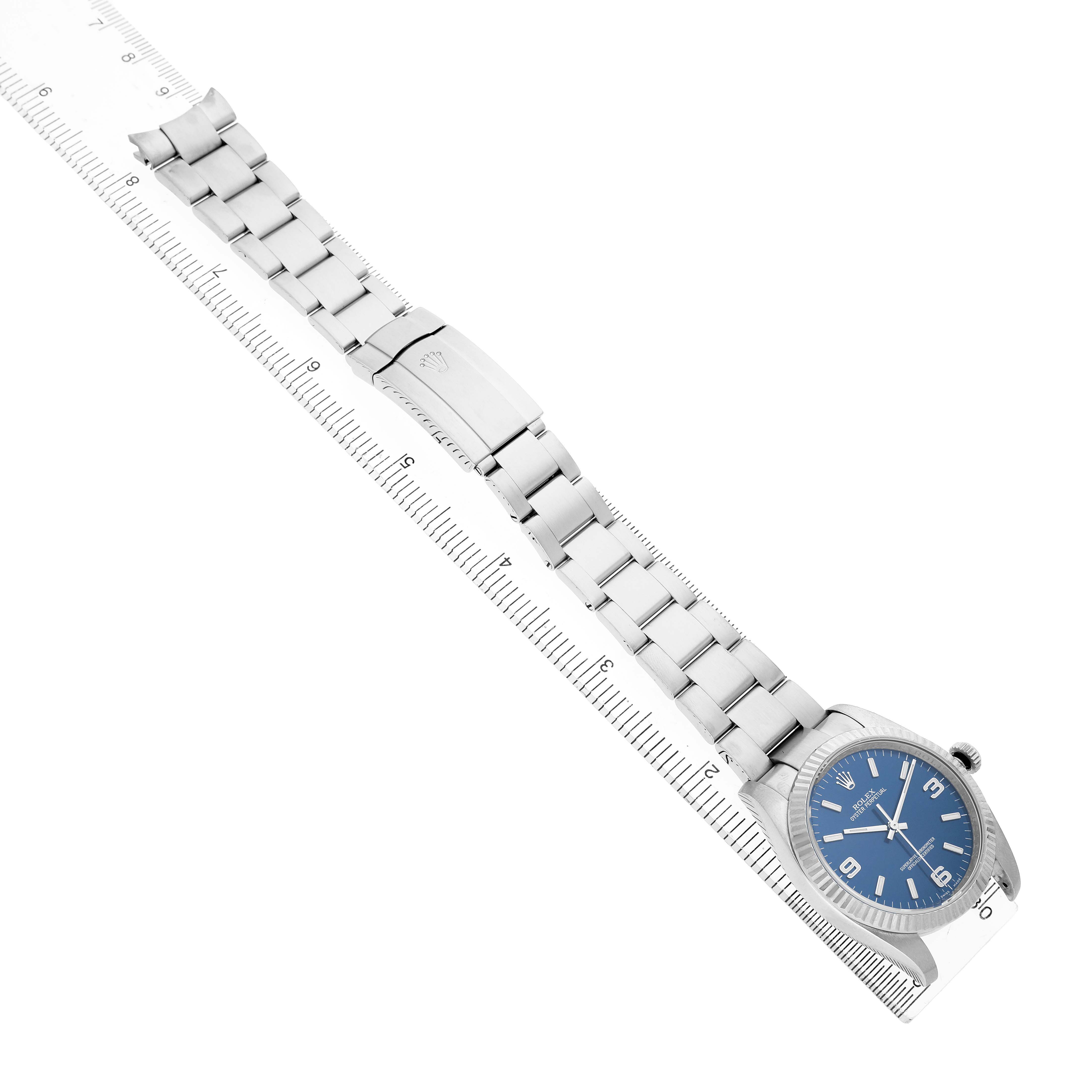 The Rolex Oyster Perpetual 116034 Men's Stainless Steel Blue Dial watch is shown diagonally, highlighting the bracelet, clasp, and blue dial face beside a ruler.