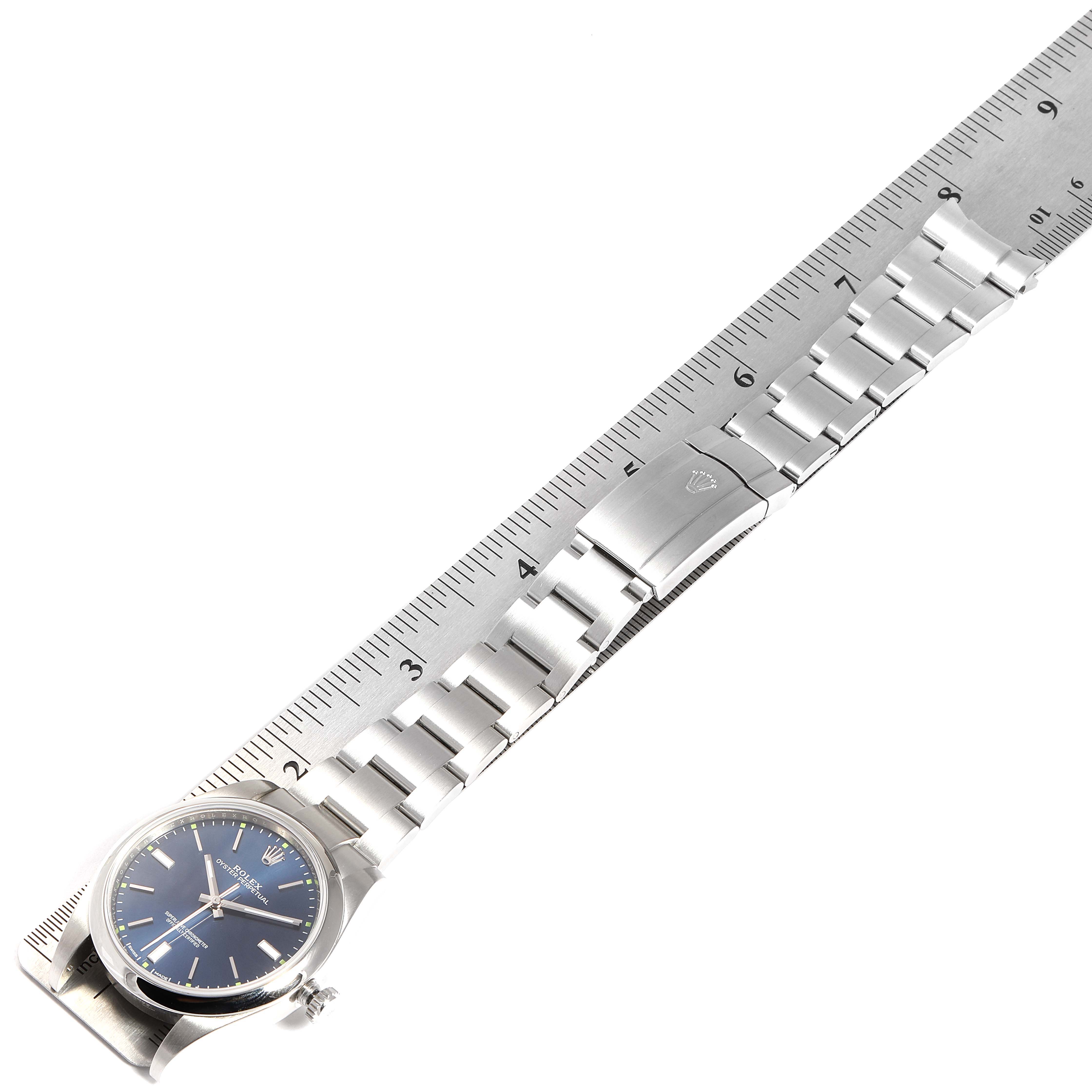 The image shows a Rolex Oyster Perpetual 114300 Men's Stainless Steel Blue Dial 114300 Men's Stainless Steel Blue Dial watch and its metal bracelet band partially extended and aligned next to a ruler.