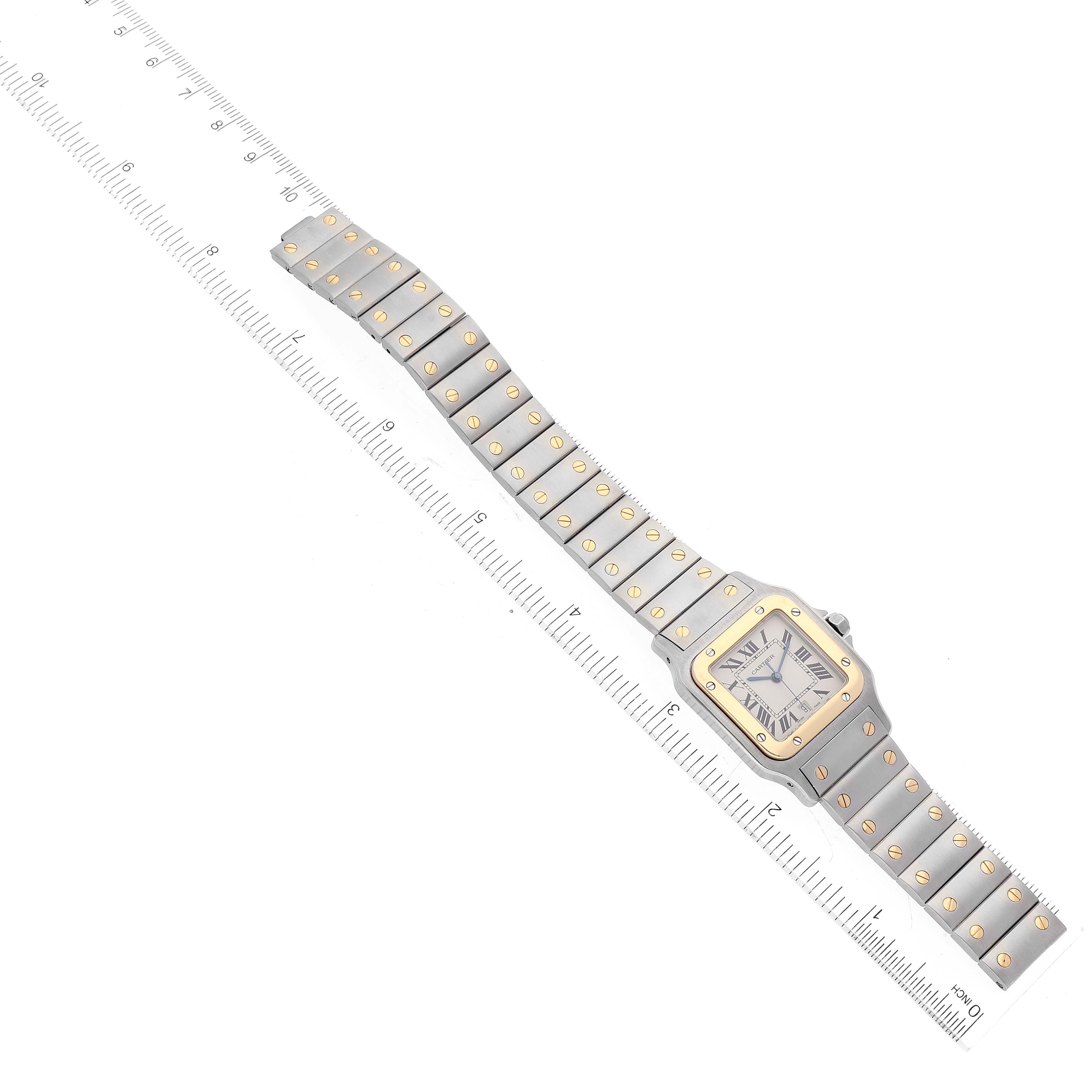 The image shows a Santos model by Cartier watch, laid flat against a ruler, displaying the watch face and full metal bracelet.