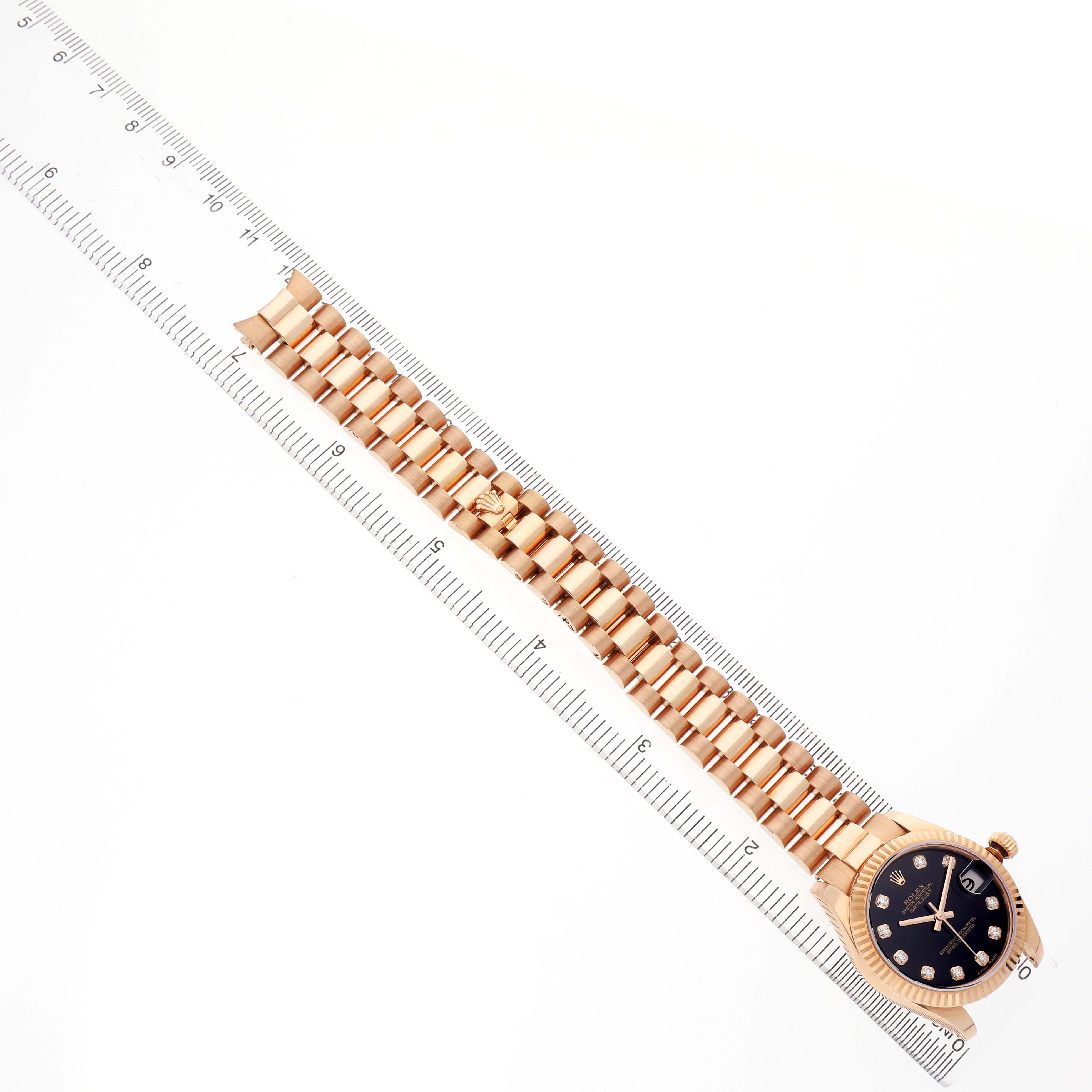 The Rolex President 178275 Women's Rose Gold Black Dial watch is shown from a top-down angle, displaying the face and band alongside a ruler for size reference.