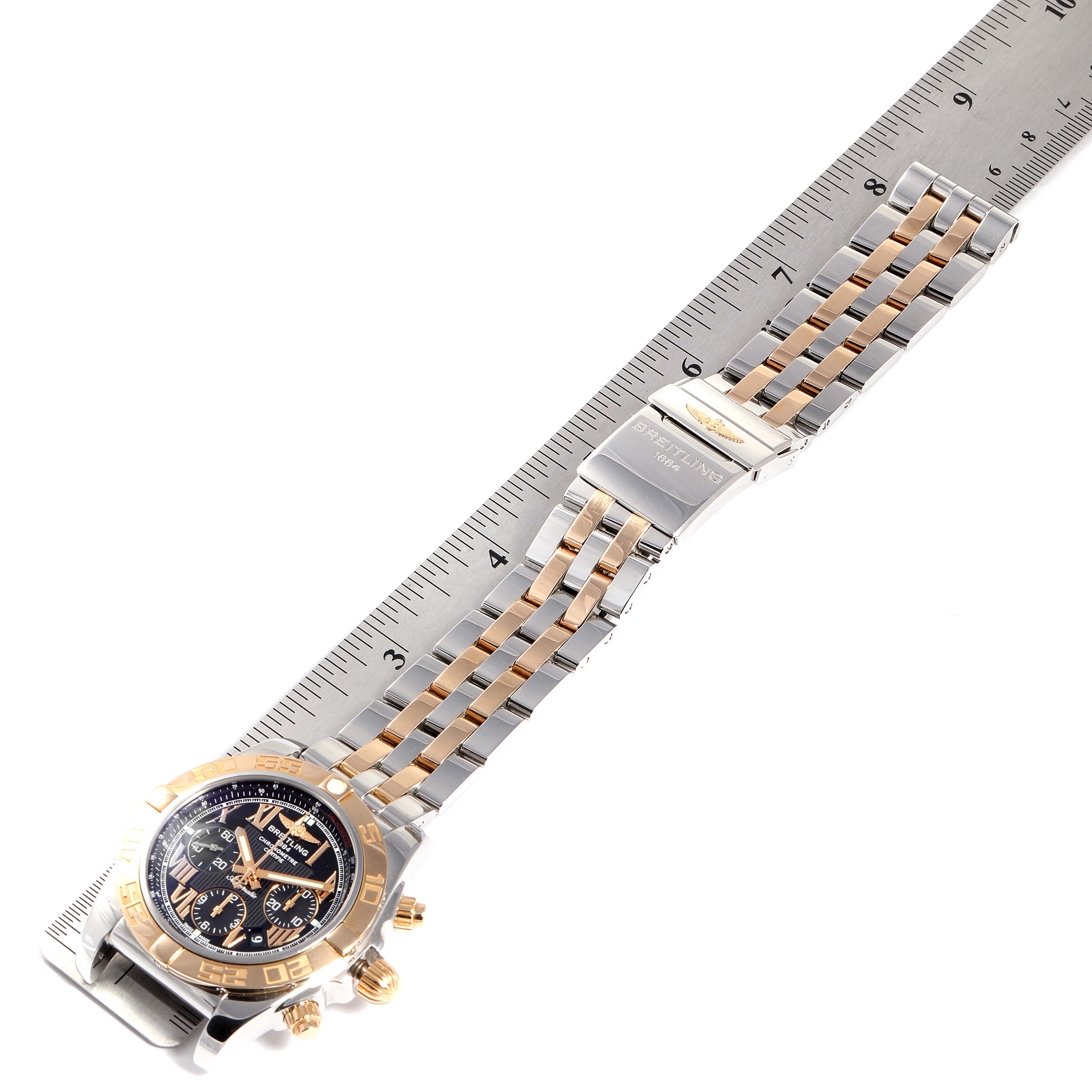 The image shows the Breitling Chronomat CB011012 Men's Steel and Gold (two tone) Black Dial CB011012 Men's Steel and Gold (two tone) Black Dial watch laid flat with a metal ruler, displaying its face, bezel, band, and clasp.