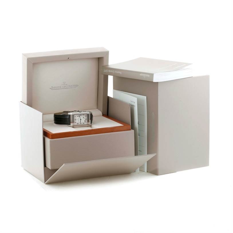 The image shows a Jaeger-LeCoultre Reverso watch from a front angle, displayed in its open box with accompanying manuals and documents.