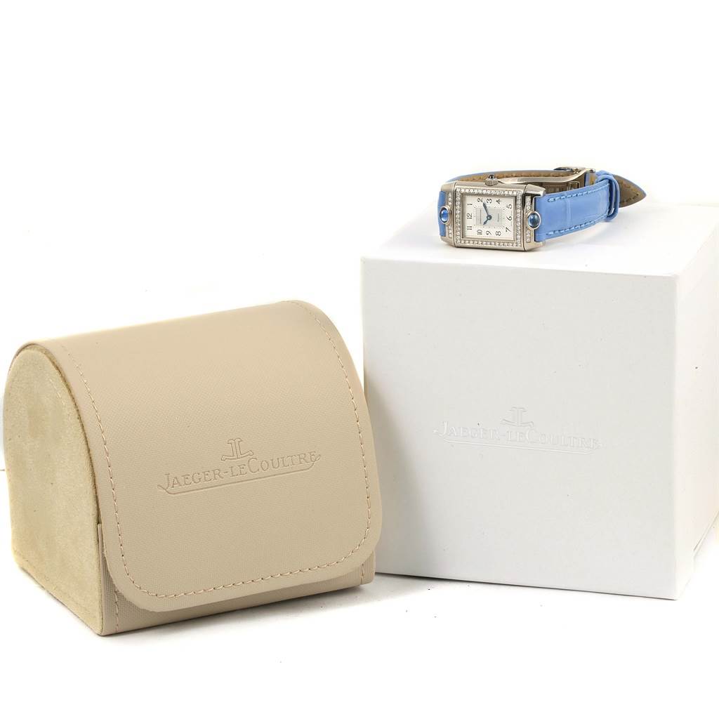 The image shows a Jaeger LeCoultre Reverso Q2623402 Women's White Gold Mother of Pearl Dial Q2623402 Women's White Gold Mother of Pearl Dial watch placed on a box with a beige travel case beside it.