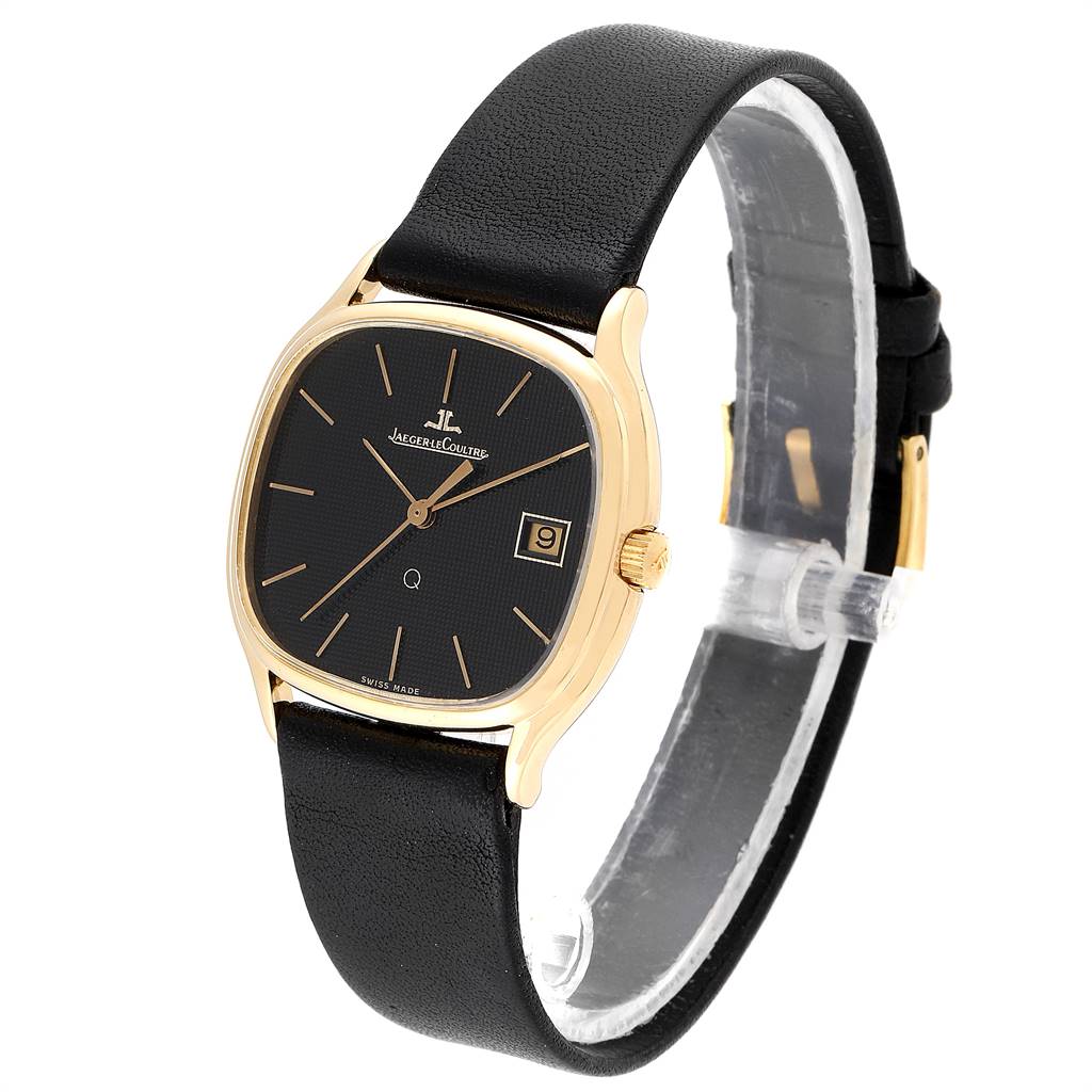 The image shows a Vintage Jaeger LeCoultre watch from a three-quarter angle, featuring its black dial, gold casing, date window, and black leather strap.
