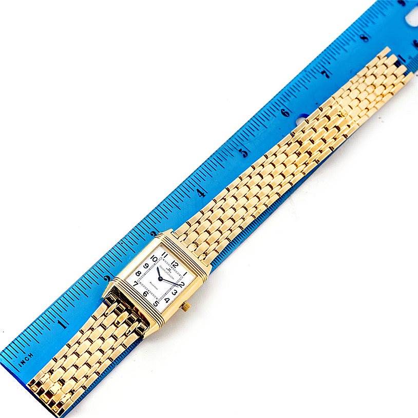 The image shows a Jaeger-LeCoultre Reverso watch with a gold rectangular face and bracelet, placed next to a blue ruler measuring its length.