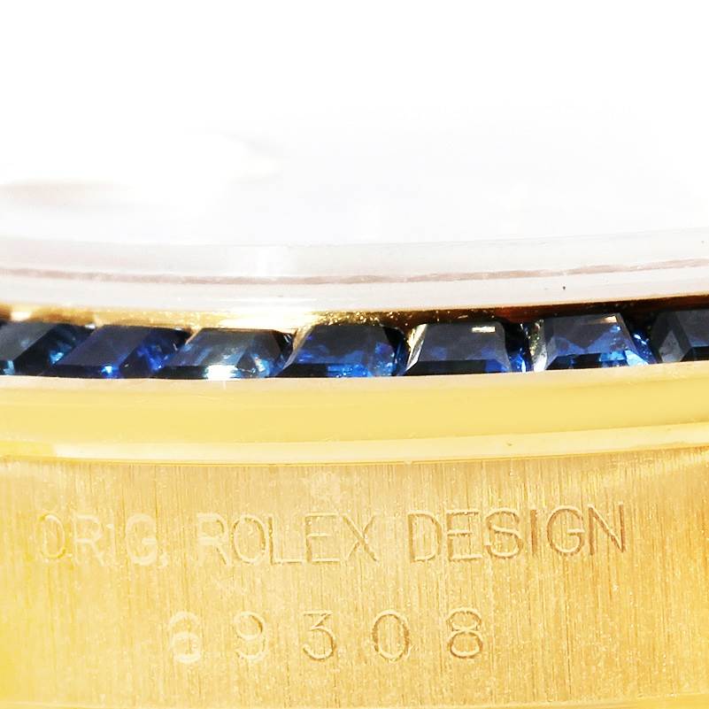 The image shows a close-up of the side of a Rolex Pearlmaster 69308 Women's Yellow Gold  69308 Women's Yellow Gold  watch, highlighting its bezel with blue stones and engraving on the case.